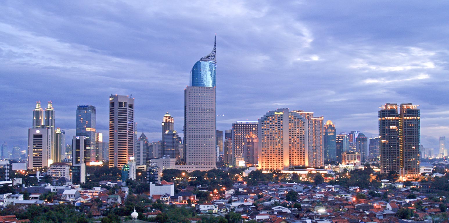 Jakarta city skyline at dusk with tall illuminated skyscrapers and residential buildings in the foreground.